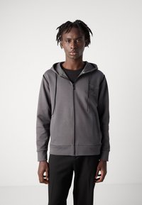 Felpa grigia con zip, dal tessuto morbido, con un accento verde sul colletto e sulle tasche laterali, indossata sopra una maglietta nera e pantaloni neri.