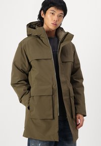 Parka imperméable vert olive avec un zip sur le devant, de grandes poches avant, une capuche ajustable et une légère brillance. Présente une coupe droite et un tissu texturé.