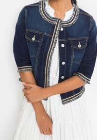 Džínová bunda tmavě modrá s přiléhavým střihem, třepícími se okraji, zlatými akcenty a knoflíkovým zapínáním; spárovaná s bílými šaty.