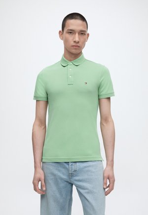 Ung mand iført en lysegrøn kortærmet polo-shirt og lyseblå jeans, stående foran en ensfarvet hvid baggrund.