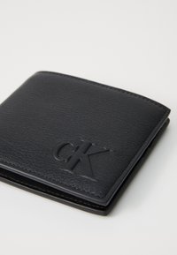 Portefeuille noir en cuir présentant une surface texturée, une forme carrée compacte, un logo "CK" en relief et une couture contrastante.