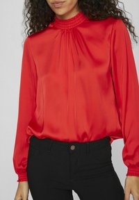 Blouse en satin rouge avec col haut smocké, longues sleeves avec des poignets élastiques, et coupe ample ; associée à un pantalon noir ajusté.