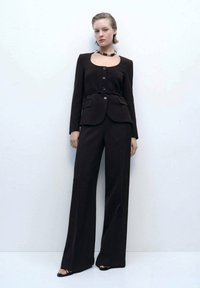 Costum negru închis, croi personalizat, cu un sacou ajustat, două buzunare și închidere cu nasturi, asortat cu pantaloni largi. Încălțăminte simplă neagră și un colier.