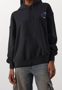 Black hooded sweatshirt with a front pocket, featuring a small graphic with "BROKEN HEART" text in blue and green on the chest. Cotton material.