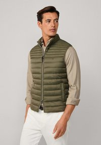 Oliivinvihreä puuvillapusero, jossa on vaakasaumat, korkea kaulus ja etuvetoketju. Yllä beige paita ja valkoiset housut.