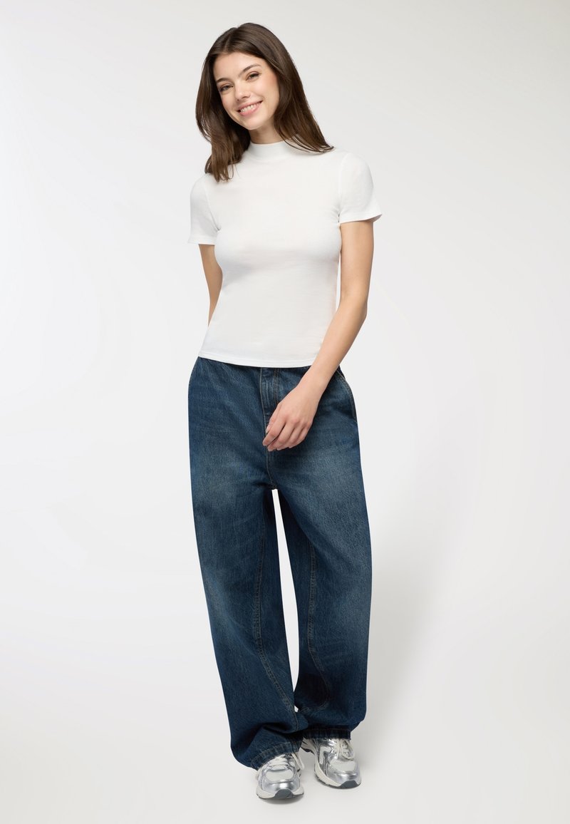 Giovane donna sorridente, che indossa un top bianco a maniche corte, jeans blu scuro larghi e sneakers argento, in piedi contro uno sfondo chiaro e uniforme.