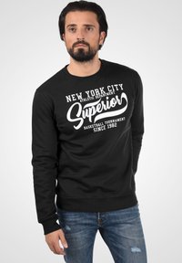 Crna dukserica s bijelim tekstom koja sadrži "New York City" i "Superior" u kurzivnom fontu, okrugli izrez i rebraste manžetne. Nosiva uz traperice.