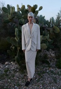 Witte pinstripe pak met een lange blazer en bijpassende rok. Gekozen in combinatie met een gemusterd hoofddoek en gouden ketting. Op de achtergrond staat een cactus.