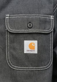 Szary materiał dżinsowy posiada przednią kieszeń z białym przeszyciem i czarnym guzikiem. Logo Carhartt jest w kolorze pomarańczowym i białym.