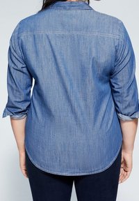 Jasnoniebieska koszula dżinsowa z zaokrąglonym dołem, podwiniętymi rękawami i szwem na plecach. Gładka tekstura, casualowy styl i kołnierz zapinany na guziki.