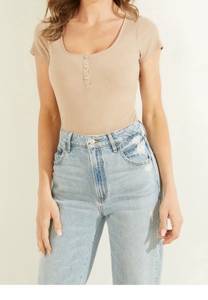 Kobieta ubrana w beżową, prążkowaną bluzkę z krótkim rękawem i guzikami, zestawioną z jasnoniebieskimi, wycieranymi jeansami z wysokim stanem.