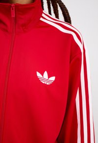 Rote Zip-Jacke mit hohem Kragen, mit weißen drei Streifen-Akzenten an den Ärmeln und einem weißen Adidas-Logo auf der linken Brust.