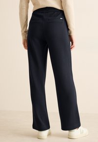 Pantalons larges marine foncé avec une surface texturée, dotés de deux poches arrière. Associés à des baskets blanches et vertes.