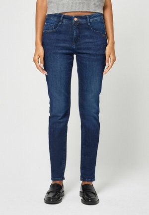 Frau trägt dunkelblaue Slim-Fit-Jeans, einen grauen kurzen Pullover und schwarze Loafers und steht vor einem einfarbigen Hintergrund.