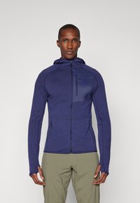Veste de sport bleu marine avec capuche, poche poitrine zippée et poches latérales. Dotée d'un tissu texturé et de passe-pouces aux poignets.