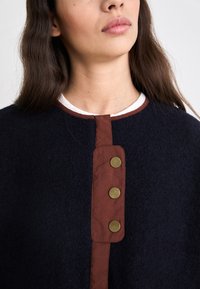 Top in misto lana navy, caratterizzato da un colletto rotondo con rifiniture bianche, completato da una patta con bottoni marroni e tre grandi bottoni in ottone.
