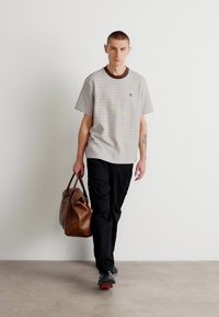 Pasiasty t-shirt z krótkim rękawem i brązowym kołnierzykiem, czarne spodnie cargo oraz brązowa skórzana torba. Model w czarnych butach, idący na neutralnym tle.