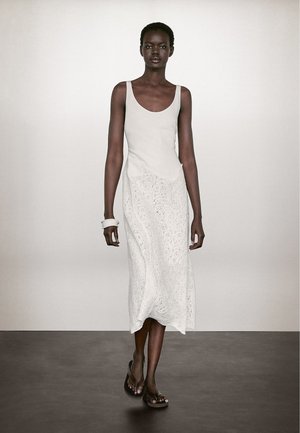 White sleeveless dress with lace texture and flowing silhouette, paired with a white bangle and brown sandals, presented on a model.