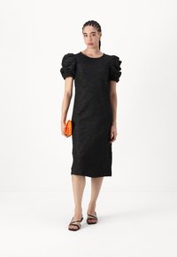 Μαύρο φόρεμα με φουσκωτά μανίκια, υφασμάτινο σχέδιο με λουλούδια, μέχρι το γόνατο, συνδυασμένο με μια ζωντανή πορτοκαλί τσάντα κλασική με παγιέτες και μαύρες σανδάλια με λουράκια.