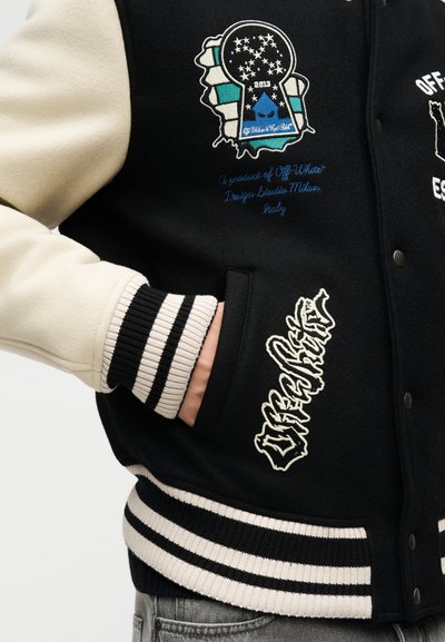 Μαύρο και κρεμ πανωφόρι varsity με κεντημένα μπαλώματα, ρίγες στις μανσέτες και ένα χέρι στην μπροστινή τσέπη.