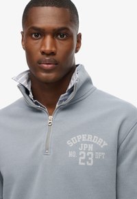 Sudadera con cremallera de color azul claro y cuello acanalado, con un forro de cuadros en contraste. "Superdry JPN No. 23 DPT" impreso en blanco en el pecho.