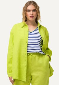 Camicia button-up verde lime con texture e vestibilità rilassata sopra una t-shirt a V con righe blu e bianche. Pantaloni abbinati verde lime.