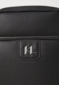 Sac en cuir noir texturé avec une fermeture éclair argentée et un patch logo en relief bien visible. Gros plan sur les matériaux et les détails de conception.