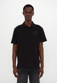Svart polo-skjorte laget av glatt stoff, med kort krage, tre-knapps lukking og et teksturert emblem på brystet.