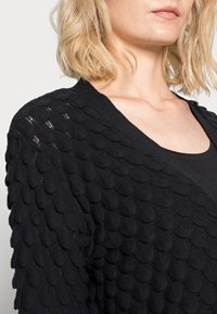 Blonde woman wearing a black textured knit garment with a scalloped pattern and a black scoop-neck top underneath.