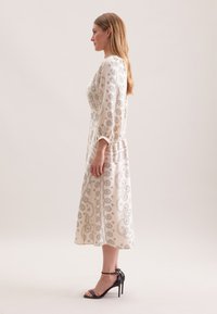 Cremefarbenes Langarmkleid mit silbernem Paisleymuster, tailliert und in Midi-Länge. Schwarze, hochhackige Sandalen runden den Look ab.
