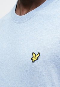 Světle modré bavlněné tričko s kulatým výstřihem, na levé straně hrudi s vyšitým černo-žlutým motivem ptáka.