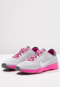 Harmaat verkkojuoksukengät, joissa on vaaleanpunaisia yksityiskohtia, valkoinen swoosh-logo, pyöreä kärki ja teksturoitu muotoilu. Nauhat vastaavat vaaleanpunaisia yksityiskohtia.