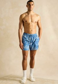 Pantaloni scurți de înot bleumarin cu un model nautic alb, talie elastică, buzunare laterale, purtați cu sosete albe până la glezne și sandale cu slidere.