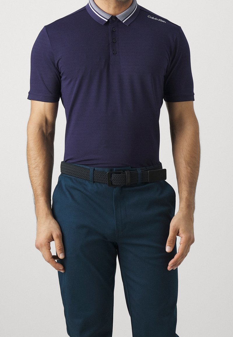 Homme portant un polo bleu marine à manches courtes avec un col rayé et un pantalon bleu foncé maintenu par une ceinture tressée noire.
