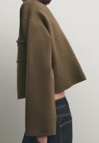 Olive green cropped jacket with wide sleeves, a textured fabric, and a front pocket with a gold button detail.