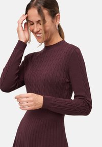 Pull en maille de couleur bordeaux foncé avec une texture côtelée et un motif en cable. Il présente des manches longues et un col rond ajusté et haut.
