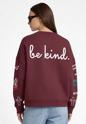 Donna con occhiali da sole che indossa una felpa bordeaux con "be kind." in grandi lettere sul retro, jeans azzurri e disegni grafici sulle maniche.