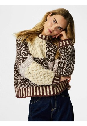 Mladá žena opierajúca si hlavu o ruku, oblečená v hnedom a krémovom pletenom svetri so vzorom a tmavých džínsoch, na jednoduchom pozadí.