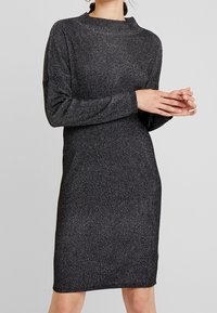 Abito a maglia nera a costine con scollatura alta e maniche lunghe, caratterizzato da una silhouette aderente e un leggero luccichio su tutta la stoffa.