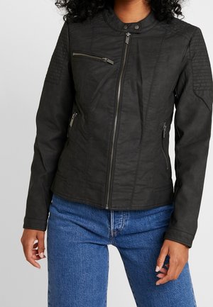 Femme portant une veste noire zippée avec des poches zippées et des empiècements matelassés aux épaules, associée à un jean bleu, debout devant un fond uni.