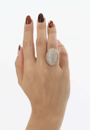 Anello ovale con fascia in oro testurizzato, incastonato con molte pietre scintillanti; indossato su una mano con unghie marroni e decorate da motivi.