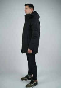 Parka noir imperméable avec capuche, présentant une coupe droite, des poches zippées et un minimum de branding, portée sur un jean foncé et des baskets vertes.