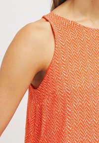 Robe sans manches orange présentant un motif en zigzag texturé blanc, avec un large décolleté et une coupe ample, visible sur l'épaule et le côté.