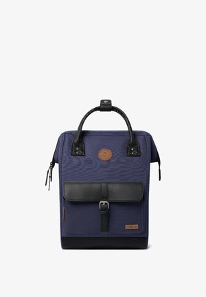 Marineblaue Rucksack mit einem strukturierten Klappdeckel aus schwarzem Leder, bronzefarbenen Beschlägen, Stoffgriffen und einem runden Logo-Aufnäher auf der Vorderseite.