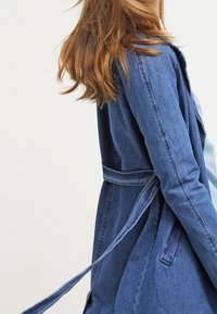 Džínová bunda ve středně modré barvě s přiléhavým střihem, dlouhými rukávy a pásem v pase. Má hladkou strukturu a rovný siluetu.