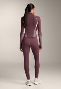 Mauve hosszú ujjú sportfelső, kontrasztos fehér díszítéssel, amelyhez illő, testhezálló leggings tartozik. Sima, rugalmas anyag és varrás nélküli dizájn részletek jellemzik.