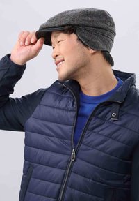 Căciulă plată cu un model herringbone în gri, accente roșii și căptușeală pentru urechi. Poartă o geacă puf în nuanță bleumarin și un pulover albastru dedesubt.