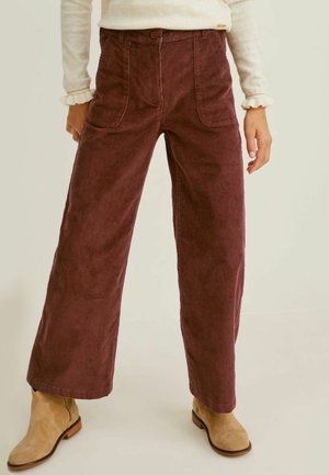 Femme portant un pantalon large en velours côtelé marron, des bottines beiges et un haut crème à manches longues avec des poignets à volants, debout devant un fond uni.