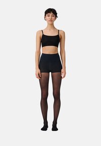 Schwarzer Bralette mit Spaghetti-Trägern, kombiniert mit hochgeschnittenen dunklen Shorts und transparenten schwarzen Strumpfhosen, die eine glatte Textur aufweisen.