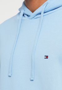 Ανοιχτό μπλε φούτερ κατασκευασμένο από μαλακό ύφασμα, διαθέτει τσέπη καγκουρό, κουκούλα με κορδόνια και ένα μικρό κεντημένο λογότυπο σε κόκκινο, λευκό και μπλε χρώμα.